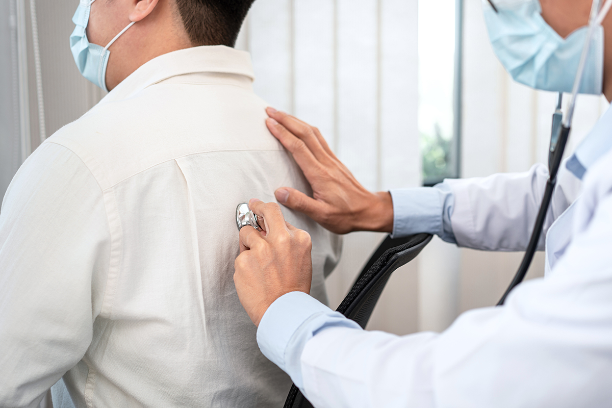 Male doctor in medical face mask using stethoscope to listening heart and lungs from male patient's back