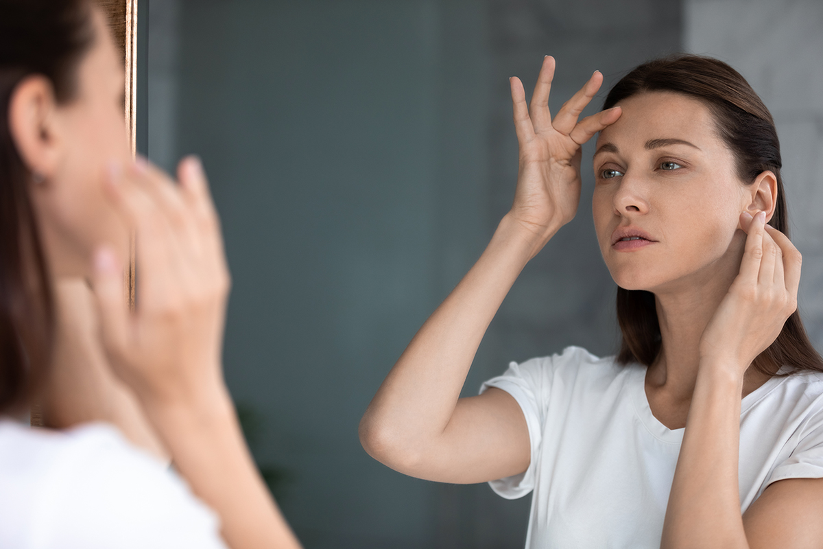 Anxious woman looking in mirror, touching forehead, confused about wrinkles