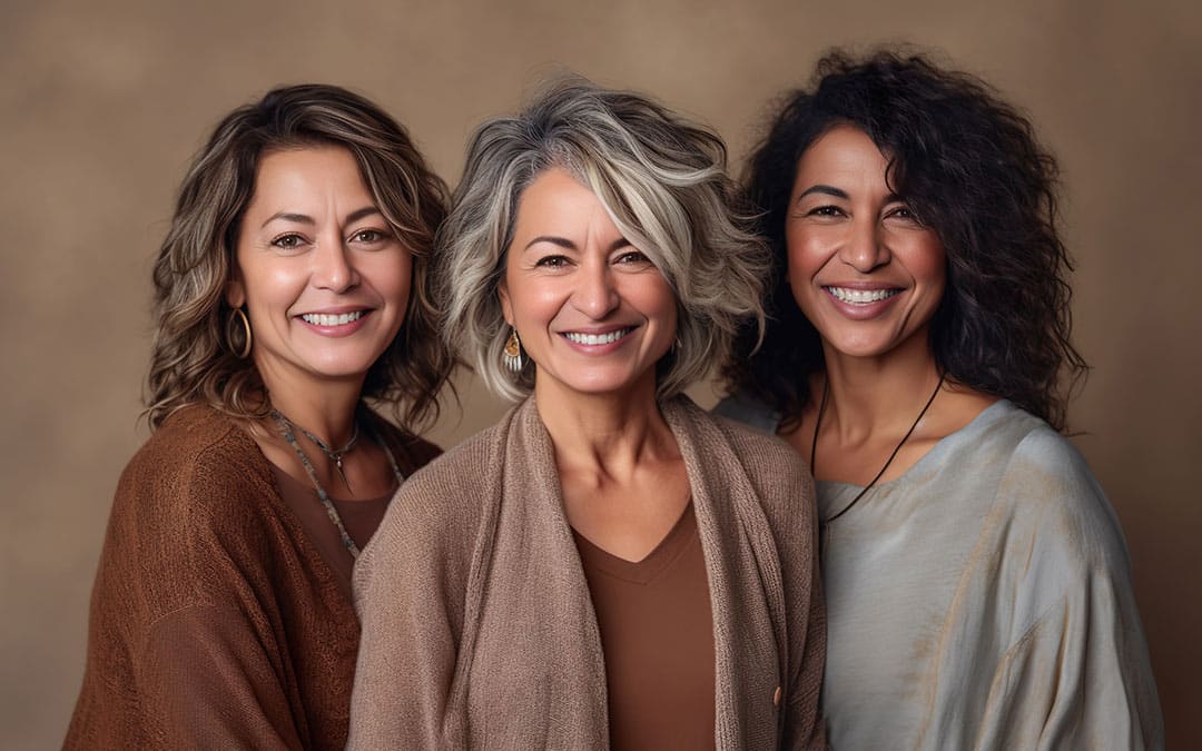 Three women of diverse ethnicities stand shoulder-to-shoulder, smiling warmly at the camera.